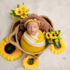 A sweet newborn wrapped in yellow and white, peacefully sleeping in a basket surrounded by bright yellow sunflowers on a fluffy white rug.
