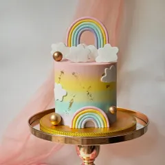 A dreamy pastel rainbow cake. This design features fondant clouds and rainbows against a soft, blended buttercream background, with gold spheres for an elegant touch.