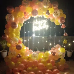 A beautiful balloon ring decoration for a terrace party at night. The yellow and pink balloons are interwoven with fairy lights, and a central "Happy Birthday" neon sign creates a warm, inviting glow.