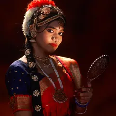 An intimate portrait of the dancer holding a traditional mirror (Aarunmula Kannadi). The lighting creates a soft glow on her face, highlighting her expression.