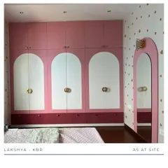 The centerpiece of this girl's room is the custom wardrobe, featuring a playful design with pink and white arched panels and unique gold hardware. The heart-patterned wallpaper adds a touch of whimsy.