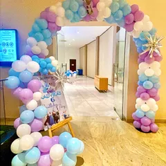 A grand balloon arch in purple, blue, and white welcomes guests to the 'Frozen' celebration. A personalized welcome sign featuring the birthday girl's name adds a special touch right at the entrance.
