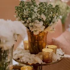 A close-up of the decor, showing simple arrangements of white flowers in textured gold vases, surrounded by candles. The petals scattered on the table add a touch of softness.