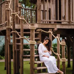 A closer shot from the same treehouse maternity session. The expecting mother is seated on the rustic wooden stairs, cradling her belly. This composition highlights the textures of the wood and rope, adding to the whimsical feel.