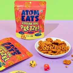 Korean BBQ pretzels styled for a game night, complete with dice and coasters. The vibrant, color-blocked background makes the scene feel energetic and fun.