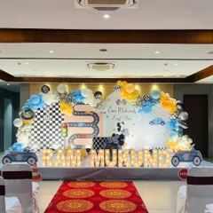 A 'Two Fast Two Curious' car-themed first birthday party. This large-scale setup features a custom printed backdrop with a racetrack, a multi-color balloon arch, car cutouts, and light-up marquee letters spelling out the birthday boy's name.