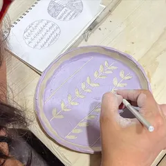 A student using a sketchbook for inspiration while carving a delicate leaf pattern onto a lavender-colored plate during a hand-building workshop.