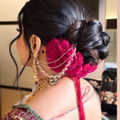 A voluminous and intricately textured bridal bun, perfect for a North Indian wedding ceremony. This style is accented with deep magenta flowers and a delicate chained accessory, creating a look that is both classic and striking from every angle.
