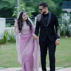 For your reception or cocktail party, we have modern and chic options. This bride wears a beautiful lavender saree with heavy silver border work, paired with a sheer dupatta. The groom complements her perfectly in a sharp black tuxedo with unique silver embroidery on the shoulders, showing our range in coordinated couple outfits beyond traditional wear.