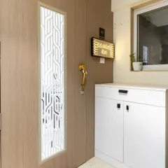 This modern entryway features a light wood door with a geometric white jali panel. The design is complemented by a functional shoe cabinet and a beautiful nameplate.