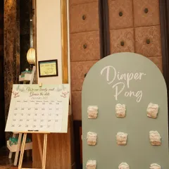 A game corner featuring 'Diaper Pong' and a 'Guess the Date' calendar. These interactive elements are designed to match the event's soft green and neutral theme.
