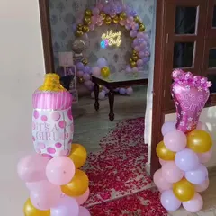 A sweet "Welcome Baby Girl" setup. The entrance features balloon pillars with "It's a Girl" foil balloons, leading to a room with a beautiful balloon arch and a "Welcome Baby" neon sign.