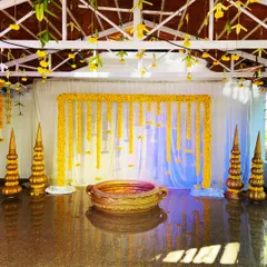 A complete setup for an outdoor Haldi ceremony. The backdrop features hanging marigold strings, and a traditional brass urli is placed in the center for the rituals, all framed by decorative golden pillars.