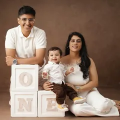 A warm family portrait with a brown backdrop, showing two mothers and their son celebrating his first birthday with 'ONE' blocks.