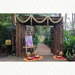 A rustic and elegant entrance for an outdoor event. The dark wood gate is adorned with jasmine garlands and flanked by brass lotuses and floral rangoli, leading guests into a serene, natural setting.