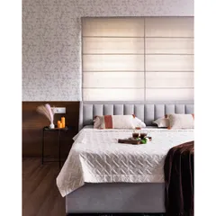 This cozy bedroom features a mix of textures for a warm and inviting feel. A soft upholstered headboard is set against a backdrop of subtle wallpaper and Roman blinds, creating a layered and comfortable space.