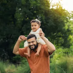 A father gives his daughter a shoulder ride during their outdoor family session. The lush green background and happy smiles make this a perfect portrait.
