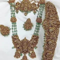 This is a magnificent antique bridal set featuring intricate carvings, goddess motifs, and green bead accents. It includes a choker, a long necklace, a statement vaddanam, and matching earrings.