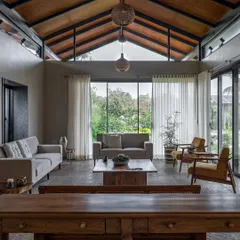 The living room of the Grove House in Alibaug features a high, pitched roof and floor-to-ceiling glass doors. This design maximizes natural light and offers expansive views of the tropical garden, creating a perfect weekend retreat for family gatherings.