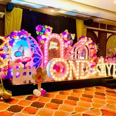 A wide view of a grand butterfly-themed first birthday party in a banquet hall. The stage is decorated with large "ONE" and name marquee letters, a light-up Ferris wheel, and intricate butterfly props.