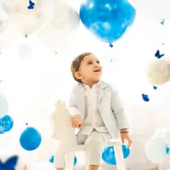 A baby boy on a rocking horse looks up in wonder at the surrounding balloons. This photo captures a moment of quiet curiosity and innocence during his first birthday celebration.