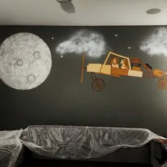 A custom wall mural we designed for a child's room. The scene features a painted night sky with a textured moon, complemented by a multi-layered airplane made from wood cutouts.