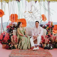 The happy couple enjoying their Mehendi ceremony, seated on a stage decorated with oversized paper flowers, colorful ribbons, and pampas grass for a bohemian-chic vibe.