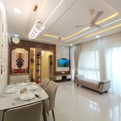 An overview of an open-plan living and dining area, showcasing how we create distinct zones within a single space. The dining set is positioned under a modern chandelier, while the living area is defined by a comfortable sofa and a dedicated TV and mandir wall. The glossy floor tiles and consistent lighting scheme tie the entire area together.