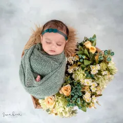 A tiny bloom nestled among flowers. This baby is wrapped in a soft green swaddle, perfectly complementing the yellow and white flowers arranged in a basket.