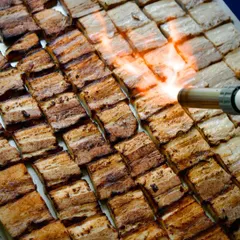 A chef uses a blowtorch to add the final char to rows of seasoned pork belly. This action shot captures the drama and precision of our live grilling stations, promising intense flavor.
