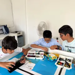 A team of three students huddles around their robot and a tablet, preparing for the IRO Techies category. They are using block-based coding to program their robot's movements and actions.