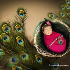 This newborn girl is styled as a divine little Radha, nestled in a basket adorned with peacock feathers. The vibrant pink wrap and serene expression create a beautiful and graceful portrait.