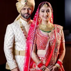 A beautiful example of a coordinated red and cream wedding outfit. The bride's red lehenga features a unique geometric pattern on the blouse, complemented by the groom's simple yet elegant cream sherwani.