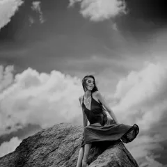 Black and white shot of a model sitting on a large boulder, with the wind catching her dress and hair