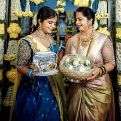 Two women at a traditional ceremony, showcasing coordinated blue and gold ensembles. Their outfits, while different in style, share a common color palette and level of intricate detail, creating a unified look.