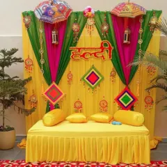 A classic Haldi ceremony backdrop designed for a home setting. This setup features a bright yellow diwan for seating, framed by vibrant yellow, pink, and green drapes, and decorated with traditional phulkari umbrellas and hanging tassels for a festive look.
