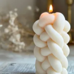The knotted weave pillar candle, shown lit to create a beautiful, soft glow. The intricate texture casts unique shadows, adding warmth and interest to your room.