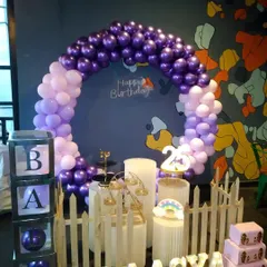 A stylish and compact birthday setup with a purple and lilac circular balloon arch. The decor is personalized with light-up marquee letters for the name "LAASYA" and includes a white picket fence and "BABY" blocks.