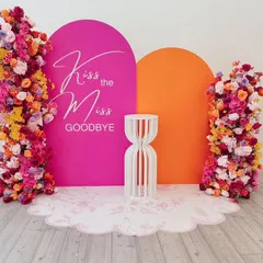 "Kiss the Miss Goodbye" with a burst of color. This vibrant setup uses a hot pink and orange backdrop with stunning floral arrangements, creating a fun and energetic photo spot.