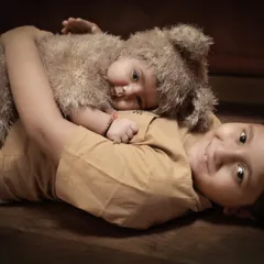 The pure love between brothers. An older brother lies on a wooden floor, gently hugging his baby sibling who is dressed in an adorable fuzzy bear costume. This is a perfect example of our sibling portraits with a newborn.