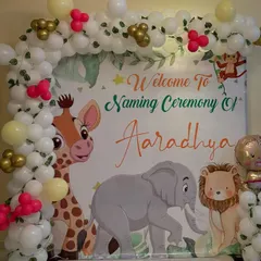 A close-up of the custom naming ceremony backdrop. The design includes cartoon animals and personalized text welcoming guests to the ceremony, surrounded by a white, gold, and red balloon garland.