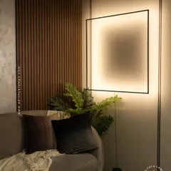We designed this suspended black frame fixture to be a piece of art in itself. It hangs from the ceiling and is grounded to the floor, creating a striking geometric statement. The light washes the wall with a soft, ambient glow, adding depth and character to this contemporary living space next to the wood-paneled wall.