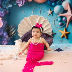 A sweet little mermaid has arrived. This six-month-old is all smiles in her bright pink mermaid outfit, sitting in a clamshell throne against a beautiful ocean-themed backdrop.