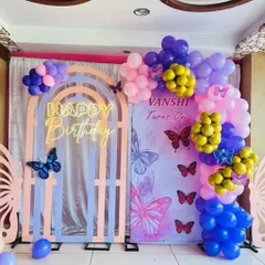 A vibrant butterfly-themed birthday party. The setup includes a custom backdrop, a colorful balloon arrangement, and a neon "Happy Birthday" sign set against a unique arched frame.
