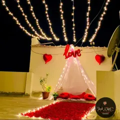 A romantic terrace decoration featuring a small canopy with a "Love" foil balloon, a rose petal pathway, and an overhead canopy of string lights.
