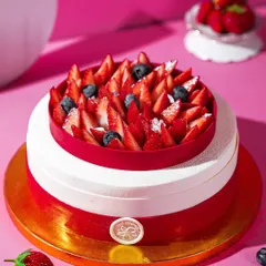 A stunning two-tiered strawberry and white chocolate mousse cake, topped with fresh strawberries and blueberries. The clean, bright styling against a pink backdrop makes this dessert look incredibly elegant.