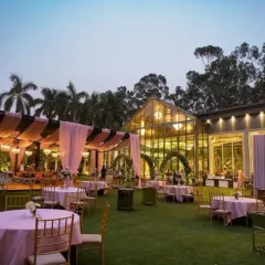 An evening reception at an outdoor lawn venue. The setup includes elegant guest seating with pink tablecloths and a beautifully lit glasshouse structure in the background.