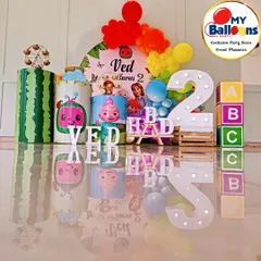 A complete Cocomelon-themed second birthday setup with a reflective floor, creating a stunning visual effect. The decor includes a rainbow balloon arch, character-printed plinths, and a large light-up number '2'.