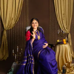 A joyful portrait of an expecting mother in a vibrant blue silk saree. She is seated on a throne-like chair in a studio decorated with golden curtains and traditional elements.