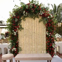 An intimate photo booth or seating area for a Nikah ceremony. The arch is decorated with red and white roses and features a backdrop of delicate white flower strings, creating a romantic and detailed corner.